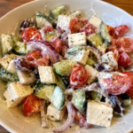 Griechischer Salat mit Feta, Gurken, Cherrytomaten, roten Zwiebeln und Kalamata-Oliven in einem cremigen Dressing, serviert in einer weißen Schüssel.